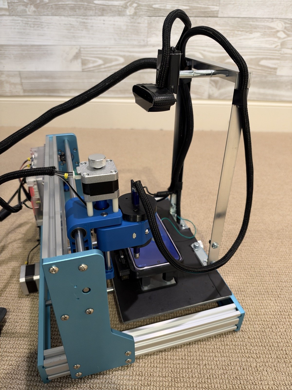 One of the two robots: a desktop CNC router with a stylus in place of its spindle, a test phone in its printed case on the bed, and a webcam looking down from the frame